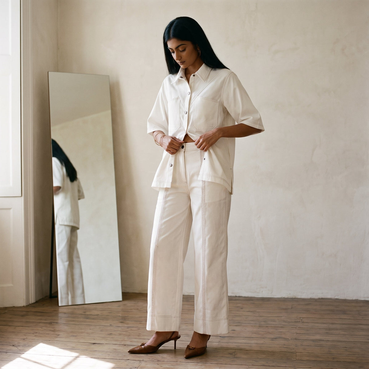 A woman in an ivory cotton co-ord set easing the trouser waistband level in front of a mirror